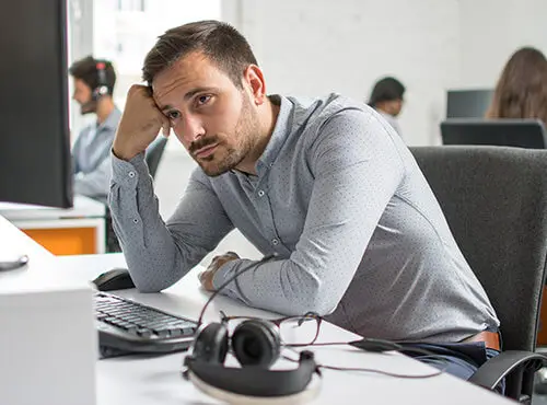En man sitter stressad och trött vid sin arbetsplats på ett kontor utan bullerskydd.